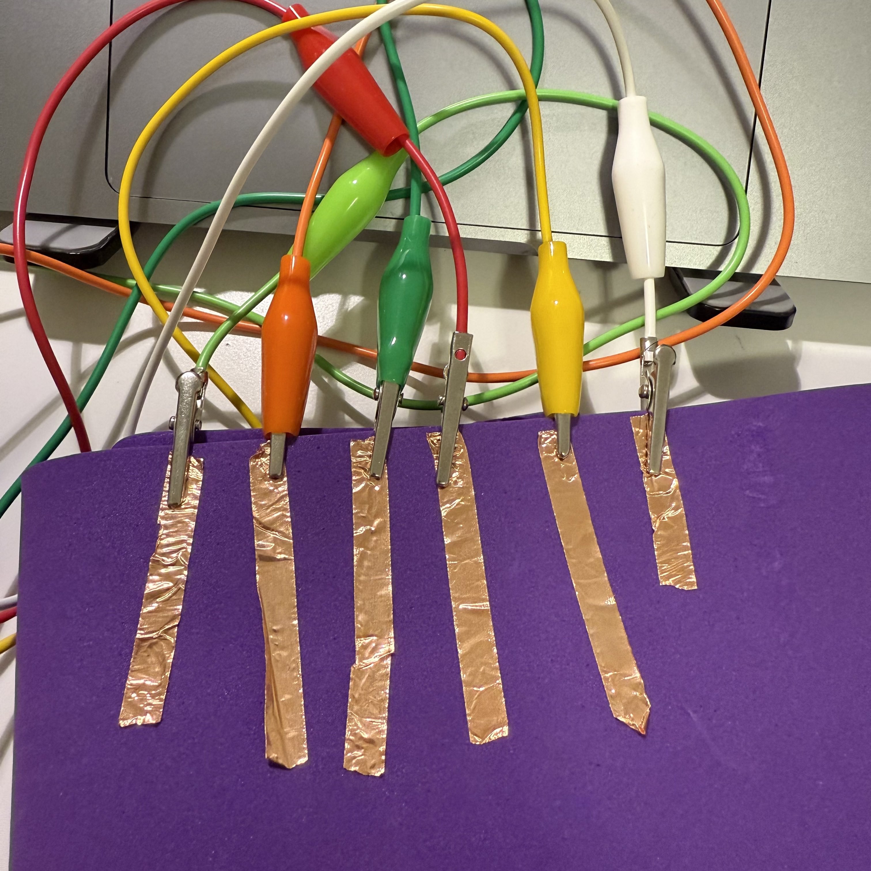 I have set up a series of copper strips on a purple board, each connected to a colorful alligator clip. This is part of my electronics project with Makey Makey, where I use these connections to turn physical touches into digital inputs.